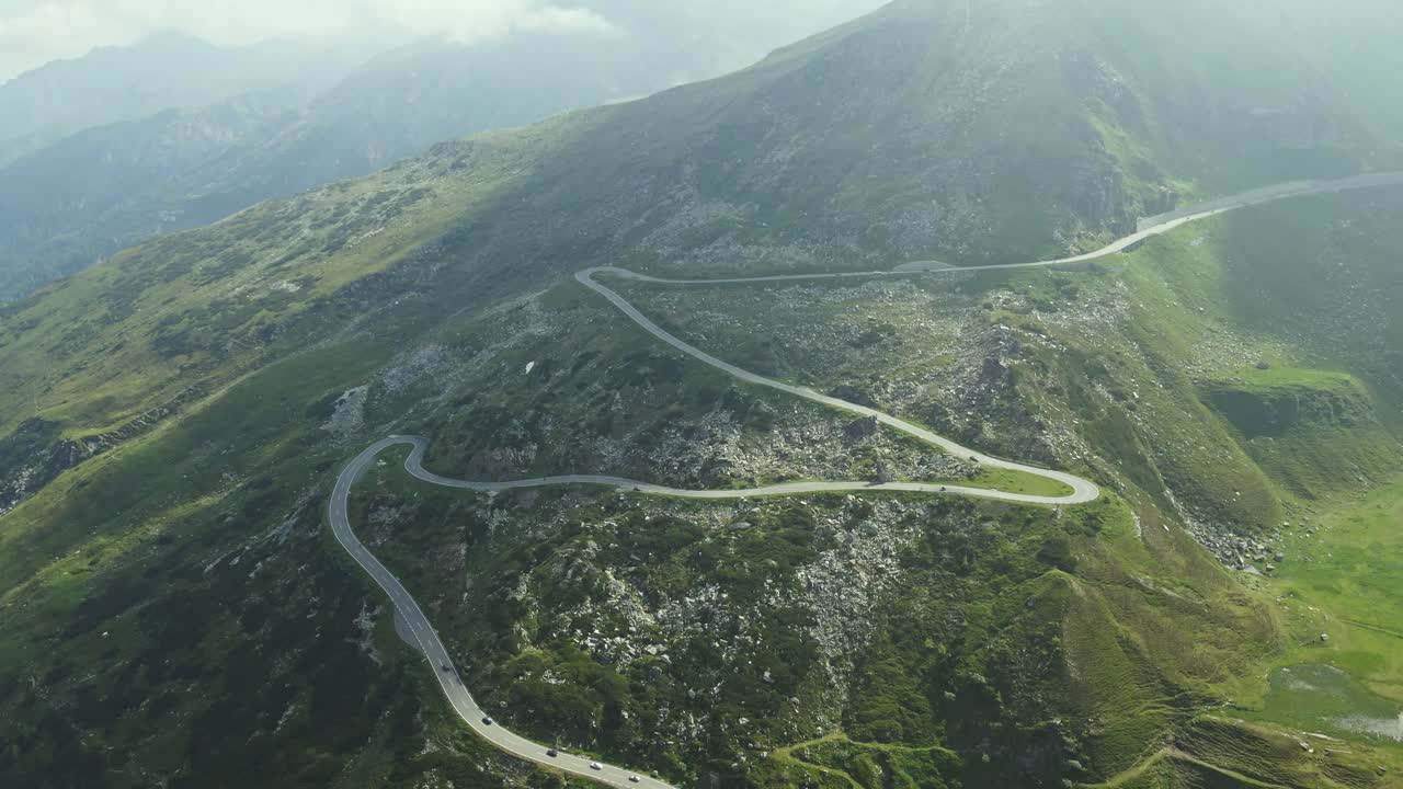 A dramatic serpentine road winds its way through a rugged mountain landscape, dotted with rocks and sparse vegetation. Several cars are visible navigating the curves.