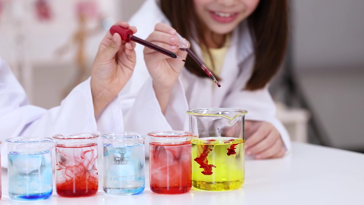 Students in white coats drop red liquid into beakers during a chemistry lesson.