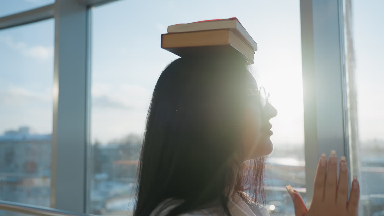 Girl balancing books on head while walking and clapping hands together near large glass windows with soft natural light illuminating snowy outdoor landscape visible through transparent mall panels