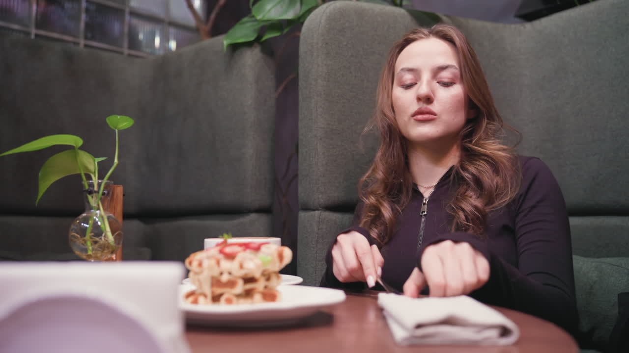 View of woman in black top with hand resting on white napkin containing cutlery on wooden table, waffles topped with strawberries and cup of coffee are placed nearby in cozy, warmly lit cafe booth