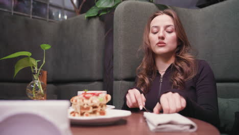 view of woman in black top with hand resting on white napkin containing cutlery on wooden table, waffles topped with strawberries and cup of coffee are placed nearby in cozy, warmly lit cafe booth