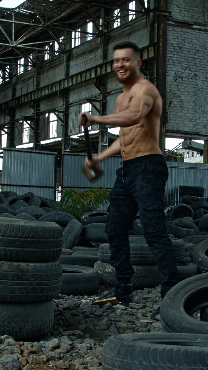 Sportsman with a heavy hammer in old plant. Sporty man trying to break pile of car tires with a metal hammer and smiles to camera. Workout in abandoned place. Vertical video