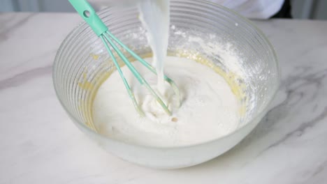 Vista-De-Cerca-De-Las-Manos-Femeninas-Vertiendo-Leche-En-El-Recipiente-Preparando-La-Masa-Y-Mezclando-Los-Ingredientes-Con-Un-Batidor.-Comida-Hecha-En-Casa