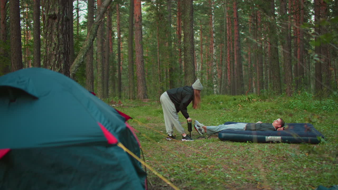 young man lies on air bed in forest clearing while woman pumps air into it beside pitched tent, surrounded by trees and daylight, capturing teamwork