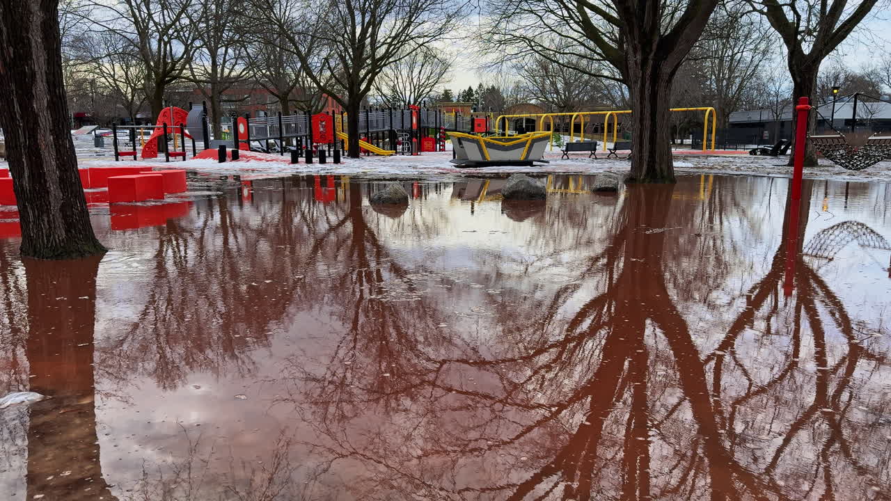 Acqua di disgelo primaverile rossastra invade un parco giochi pubblico, dove i riflessi degli alberi spogli e le strutture di gioco sommerse rivelano una scena di parco improvvisa e inutilizzabile.