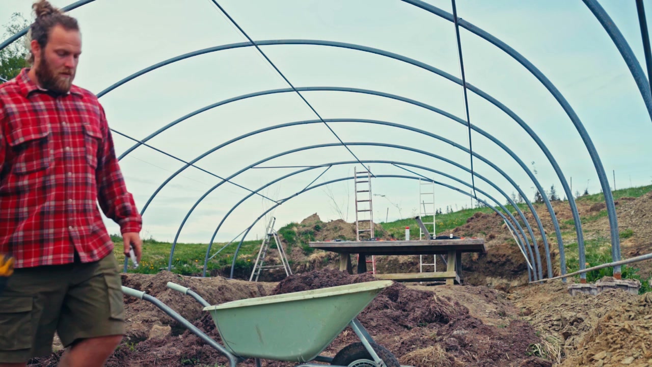 Un hombre trabaja al aire libre en la estructura de acero de un invernadero DIY en una zona de cultivo, ensamblando y trabajando en la estructura metálica. Timelapse