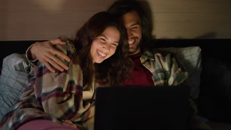 una pareja feliz viendo una película con luces apagadas en un remolque fuera de la ciudad en verano. una chica morena feliz con una camisa a cuadros rosas se acuesta con su novio moreno con una camiseta a cuadros verdes, que la abraza mientras ven una película juntos en una computadora portátil en un campamento en un trailer en la noche de verano