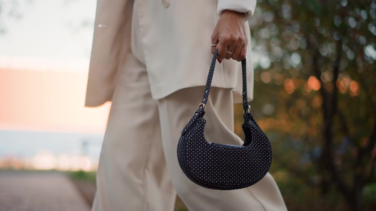 Donna che cammina all'aperto al tramonto, tenendo una borsa a mezzaluna nera con perline per il manico, con passo rilassato, un leggero ondeggiare del braccio, un outfit elegante, acqua calda e luminosa e una luce serale soffusa.