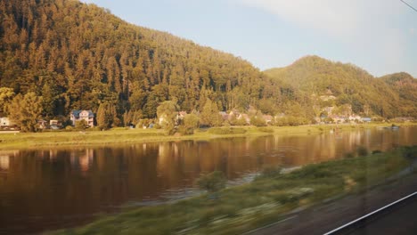 Paisaje-De-árboles-Otoñales-Junto-A-Las-Montañas-Con-Un-Río-Tranquilo-Desde-Un-Tren-En-Movimiento-En-Praga,-República-Checa