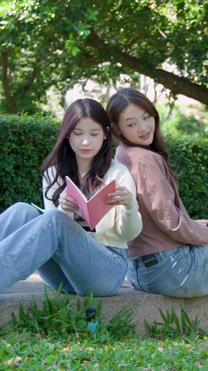 2人の若い女性が石の縁に背中合わせに座り、ノートパソコンで勉強している。