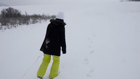 Back-view-of-female-in-winter-clothing-dragging-wooden-sleigh-behind-her-in-hilly-snowy-winter-landscape-right-pan