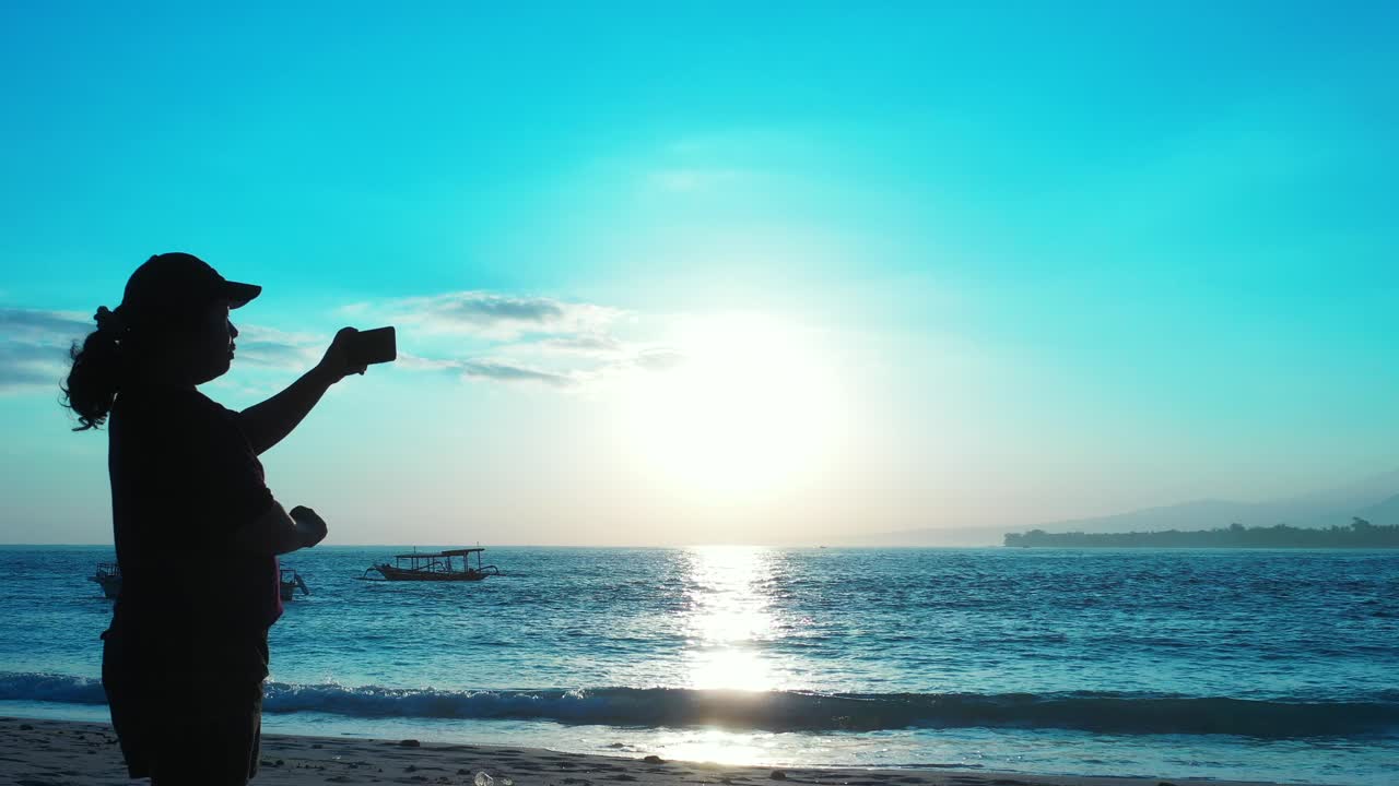 A woman alone on a shoreline of tropical beach taking photos of the beautiful afternoon sun setting over the blue water