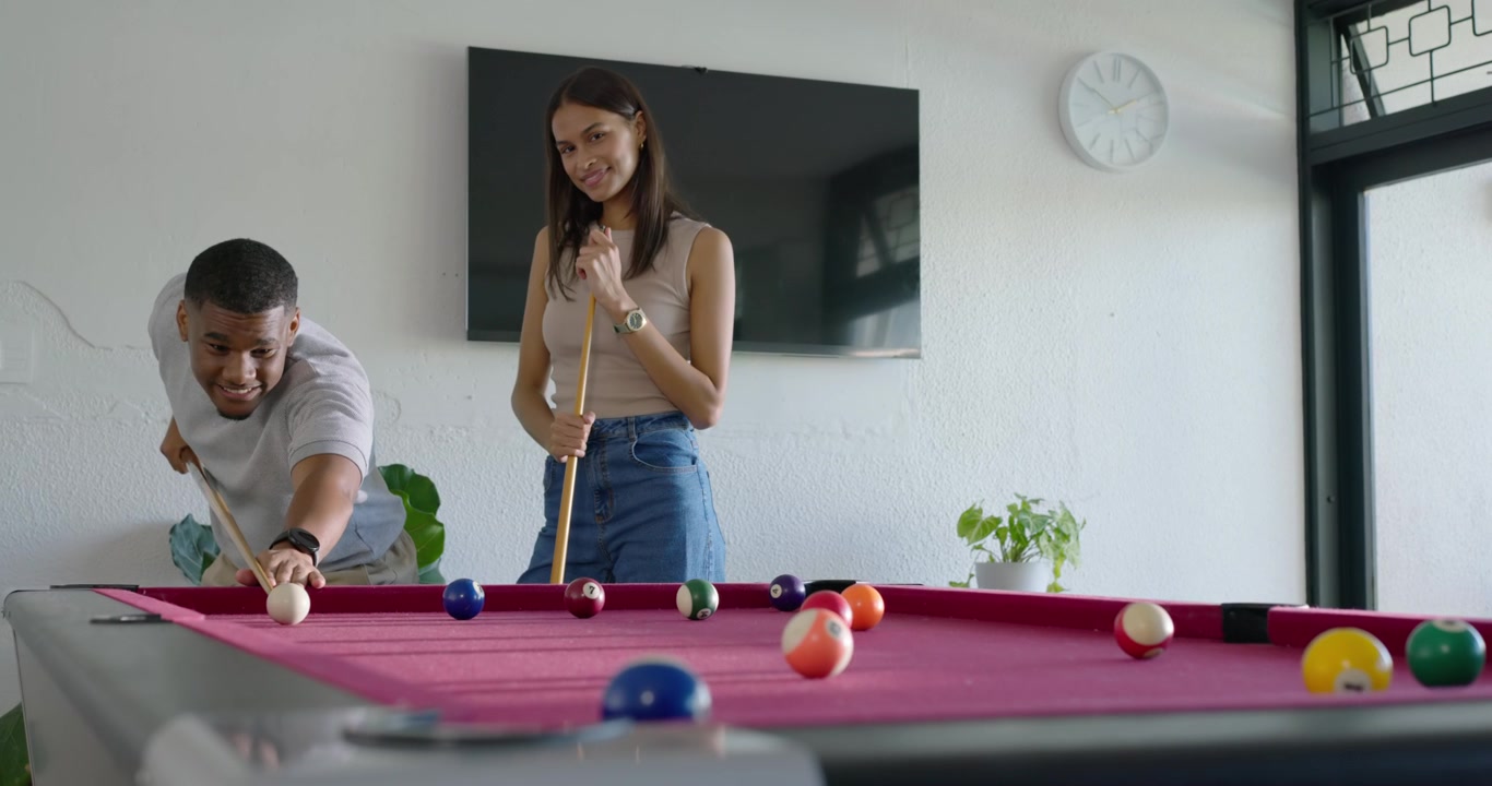 African American couple playing by pool table at home, man pointing and teaching then taking shots