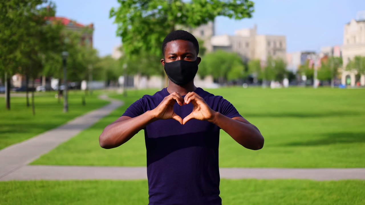 Masked Student In Park Making Heart With Hands On Green Campus Lawn, Wearing Black Face Mask And Navy Shirt,