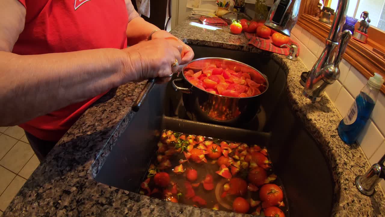 mujer mayor de pie en el fregadero de la cocina mientras prepara tomates cultivados en el jardín para enlatar