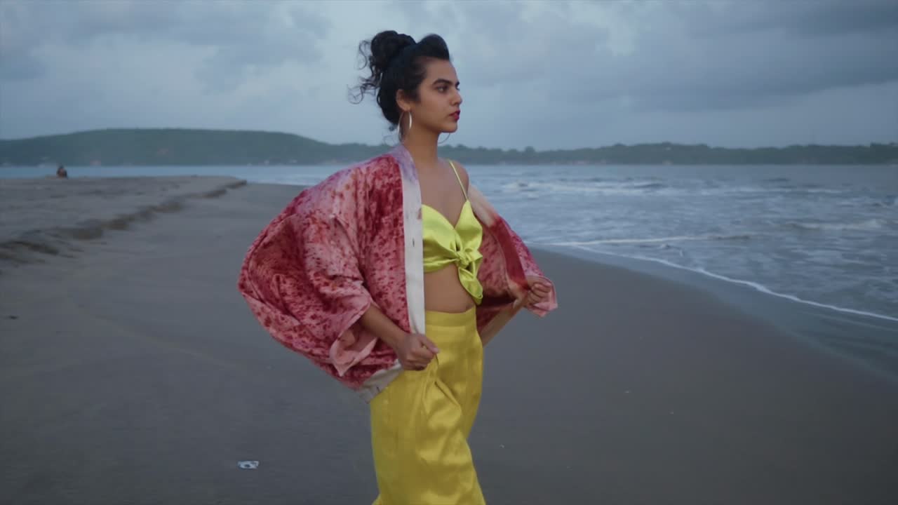 imágenes cinematográficas de una modelo de moda india con un traje amarillo y una chaqueta de seda roja en una playa de arena en el océano en un día nublado en goa india, cámara lenta, slomo