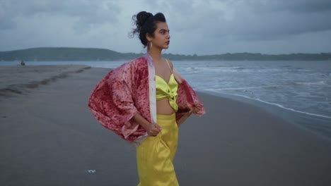 Cinematic-footage-of-an-Indian-fashion-model-wearing-a-yellow-outfit-and-a-red-silk-jacket-on-a-sandy-beach-at-the-ocean-on-a-cloudy-day-in-Goa-India,-Slow-Motion,-Slomo