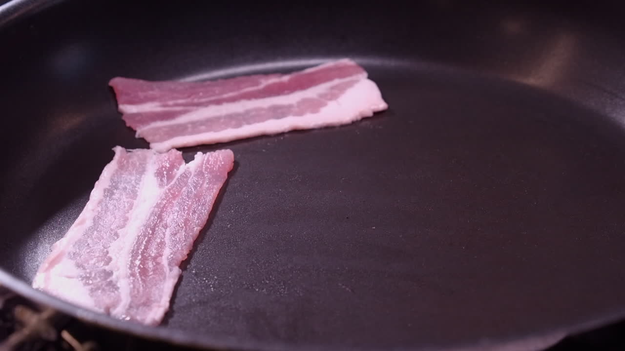 Raw bacon strips are placed into fry pan by Caucasian right hand