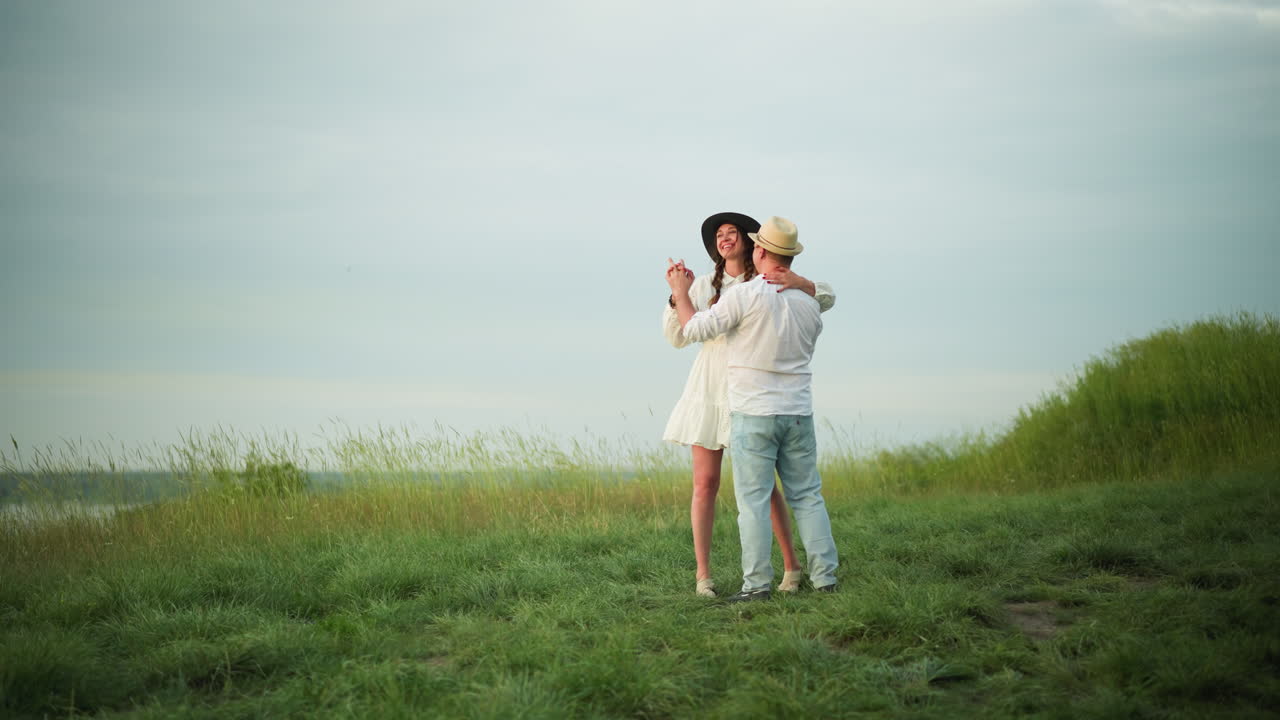 사랑하는 커플이 잔디 인 언덕에서 함께 춤을 추고, 남자는  셔츠, 모자, 청바지를 입고, 여자는  드레스와 검은 모자를 입고 있습니다. 이 장면은 부드럽고 즐거운 순간을 포착합니다.