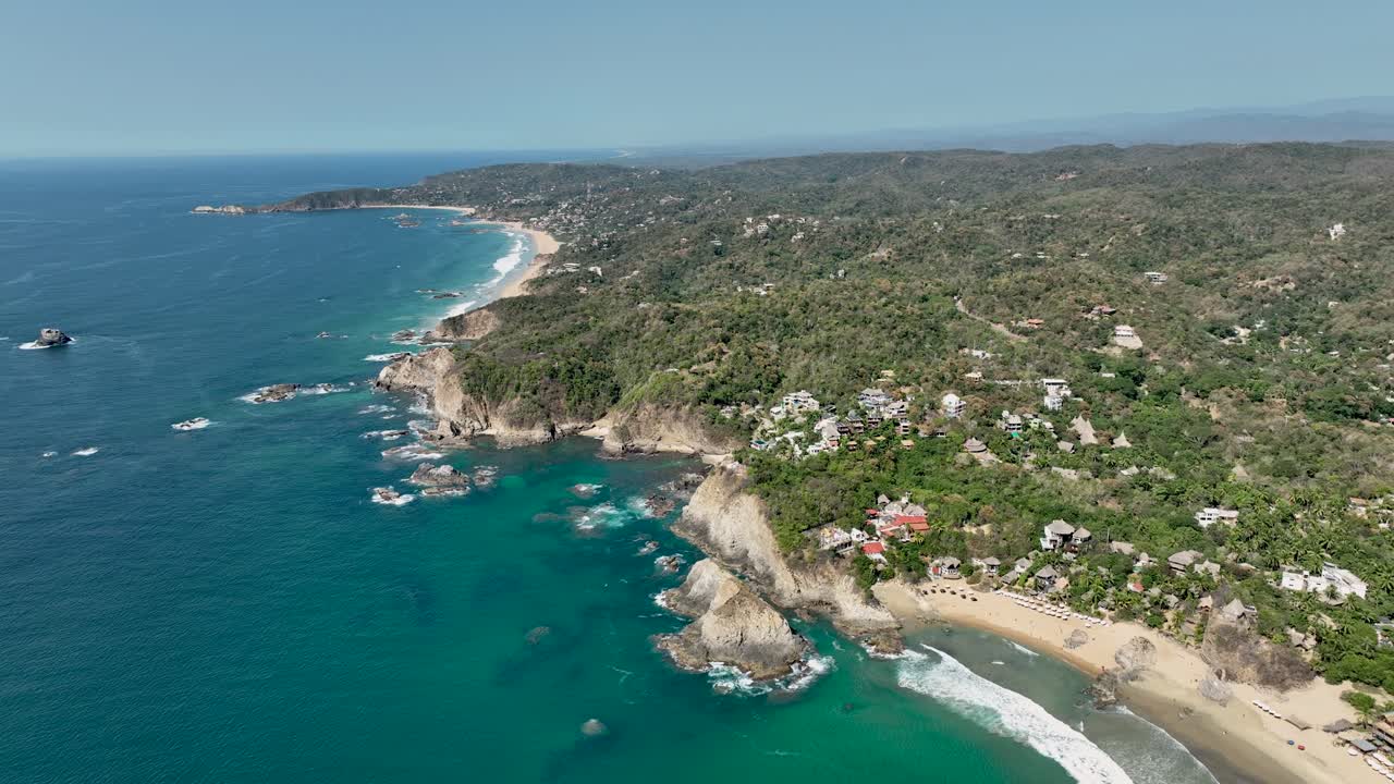 Aerial orbit around Mazunte on Mexico’s Pacific coast, revealing green hilltops, rocky cliffs and sandy coves. Clear day with expansive coastal relief; Punta Cometa visible in the distance