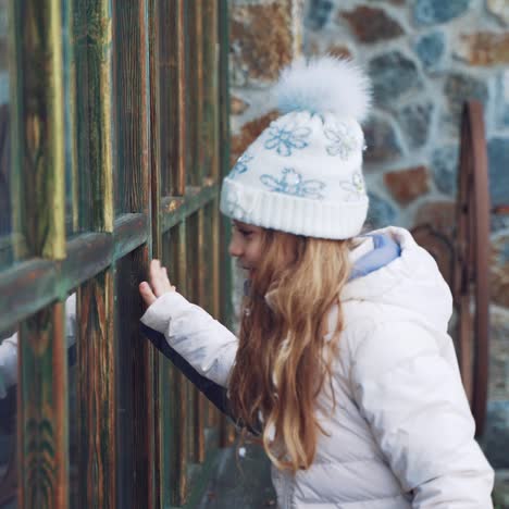 cheerful little girl with long hair and in warm clothes is peeping through the window from the street behind the elves in the winter against the background of the building. good new year spirit