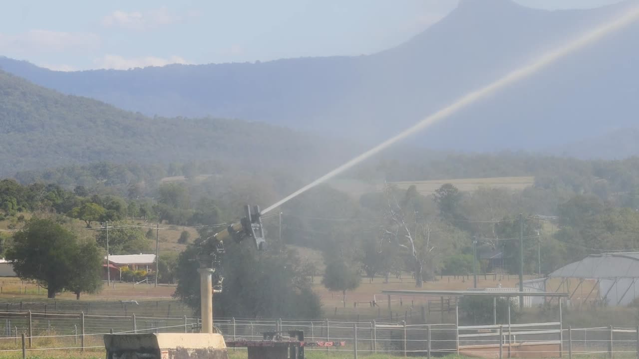 A rotating sprinkler system waters a farm field, set against a scenic mountain landscape.