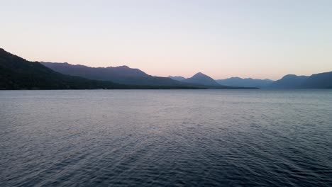 dolly out of a lake with mountains on the horizon in the blue hour and birds flying around - aerial
