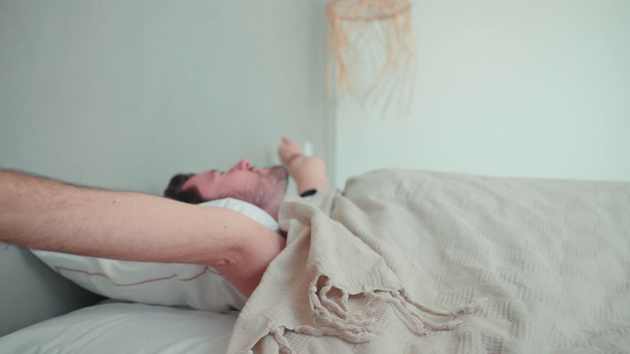 Uomo addormentato, caucasico con la barba, si sveglia al mattino, si stira stancamente, apre gli occhi, si alza rilassato nella camera da letto di casa