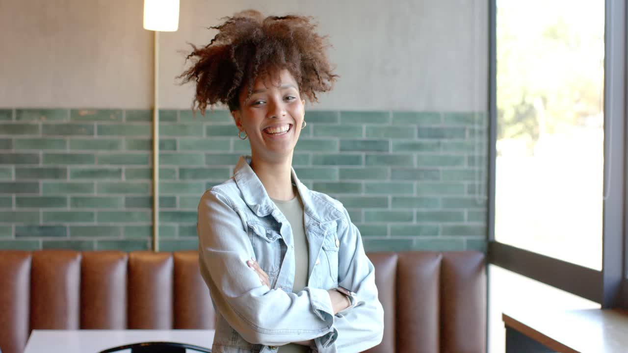 Mujer afroamericana de unos 20 años posando con las manos en las caderas en una cabina de cafetería, ajustándose la chaqueta y riendo para la cámara