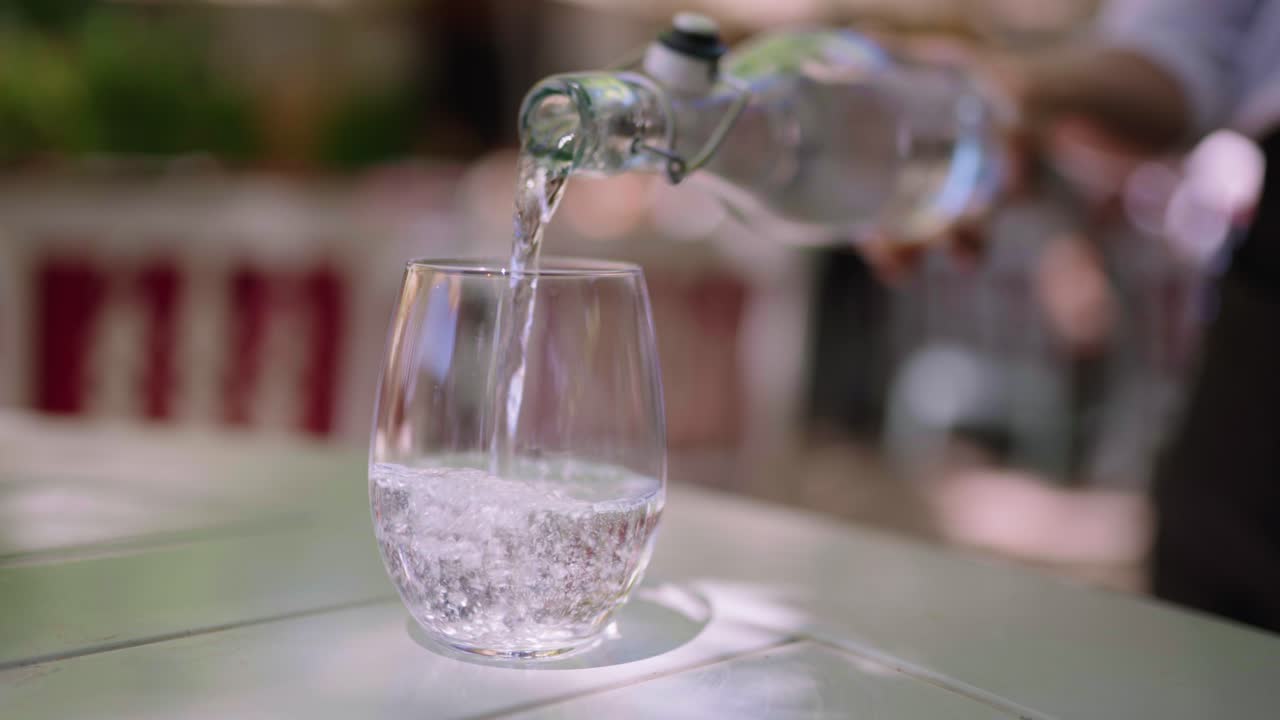 Close up clip of sparkling water being poured from a glass bottle into a drinking glass at a table setting
