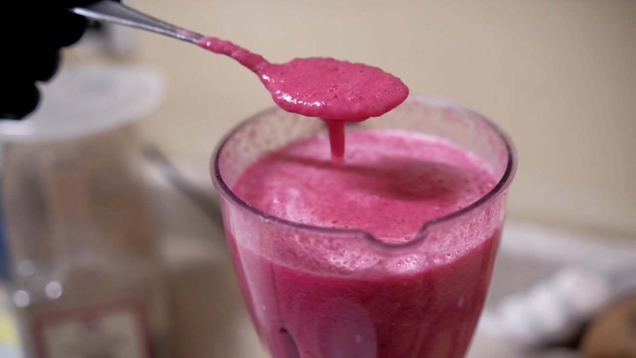 Nærbilde av en skje som løfter tykk, nyblandet rosa aguachile-saus over en blenderkopp mens en kokk sjekker den glatte teksturen og konsistensen før sjømaten anrettes.