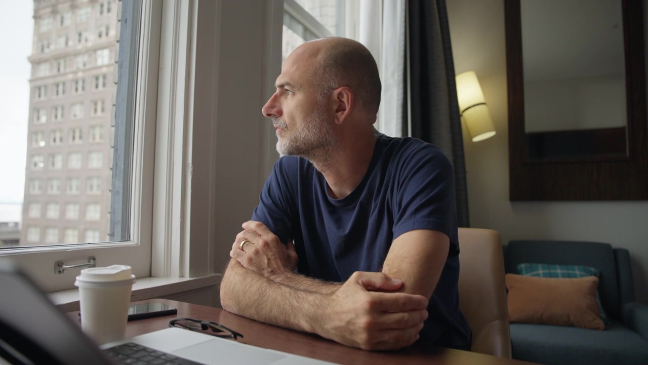 Un uomo lavora al laptop nel suo ufficio domestico, si toglie gli occhiali e guarda fuori dalla finestra con una tazza di caffè sulla scrivania