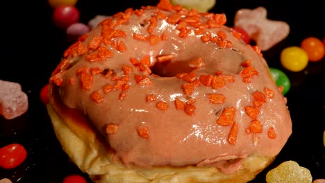 pink funny surprised glazed donut with sprinkles, striped caramel candies, colorful dragee with raisins or peanuts inside on black background