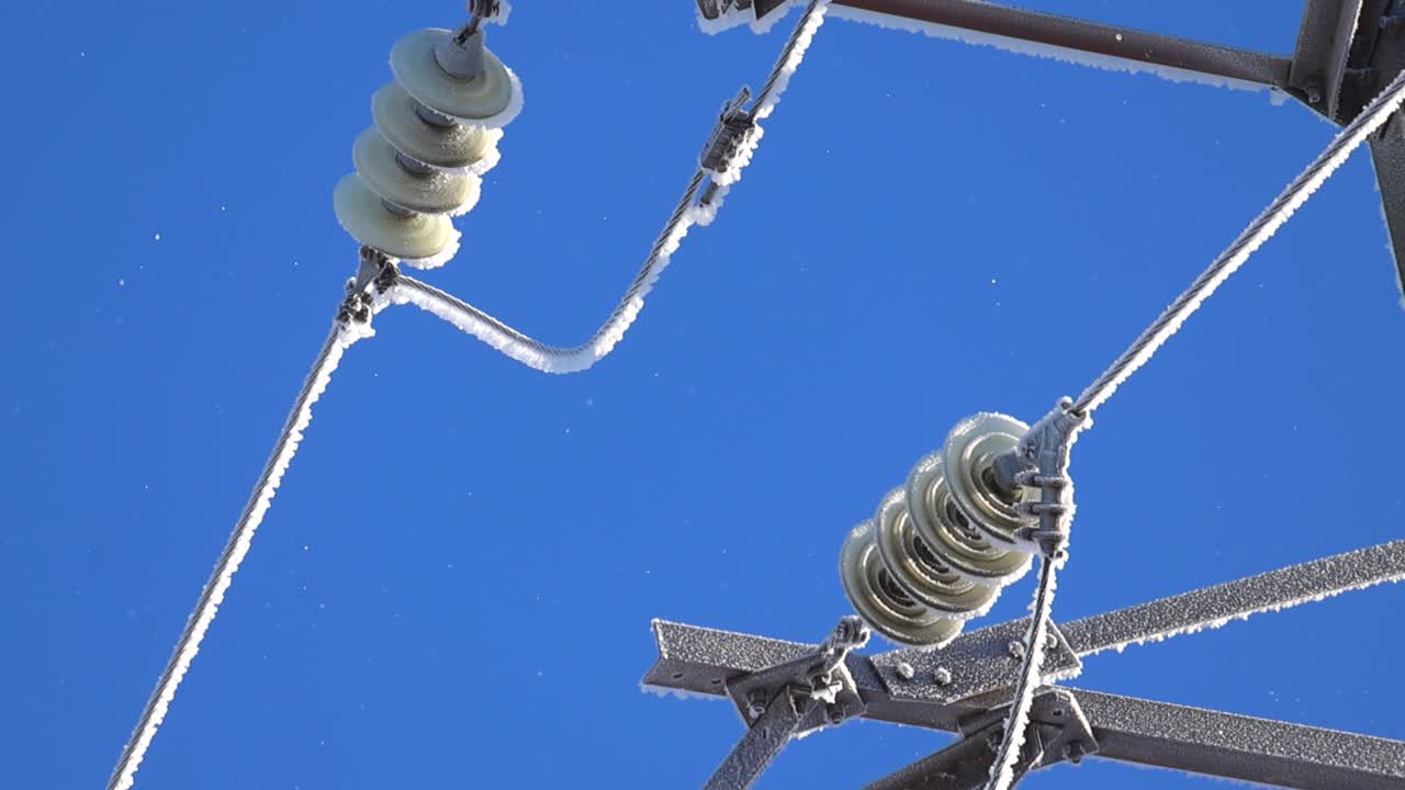 Close up ground shot reveals frozen high voltage insulators and cables on a steel pylon, with hoarfrost and ice sparkling in sunlight against a deep blue winter sky