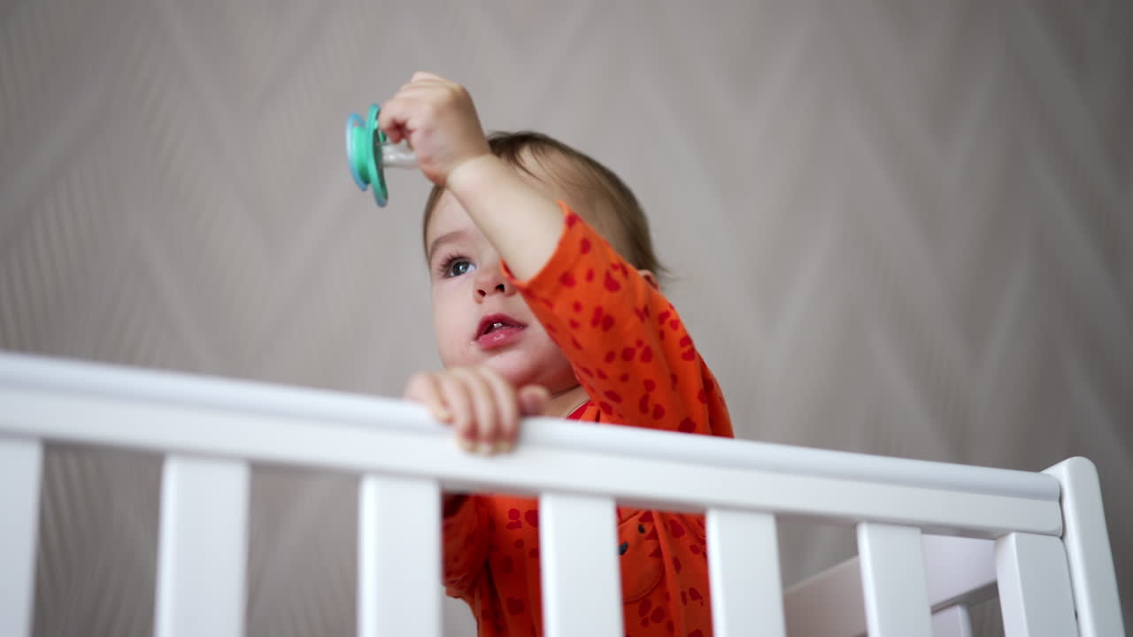 Adorable toddler boy playing with pacifier standing in the cot. Baby sits and disappears in the crib. Low angle view.