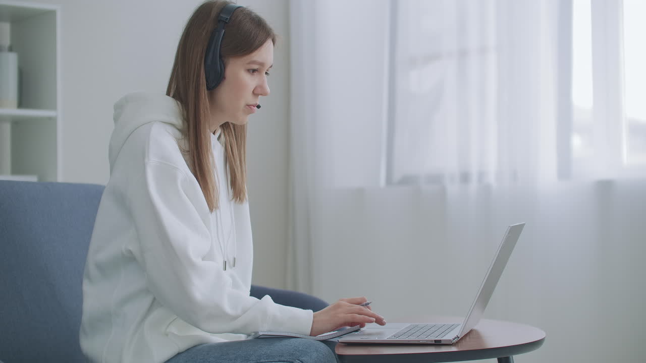 젊은 여성이 헤드 ⁇ 을 착용하여 노트북으로 회의 통화를 하며 온라인 교사와 대화를 나누고 집에서 공부하고 있습니다. 여성 학생은 컴퓨터 웹캠 채팅을 사용하여 학습하고 메모를 합니다. 원격 교육 개념