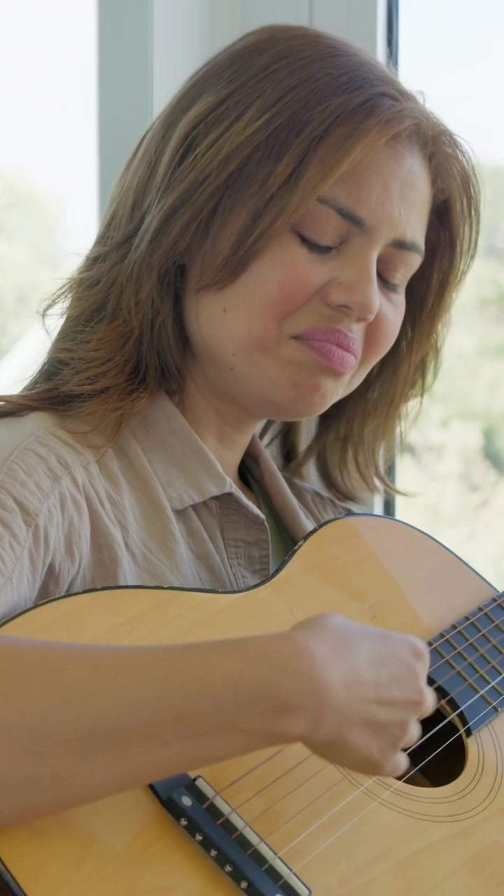 Vertical video: Cutting shot, woman strumming guitar and singing by window in tan shirt practicing