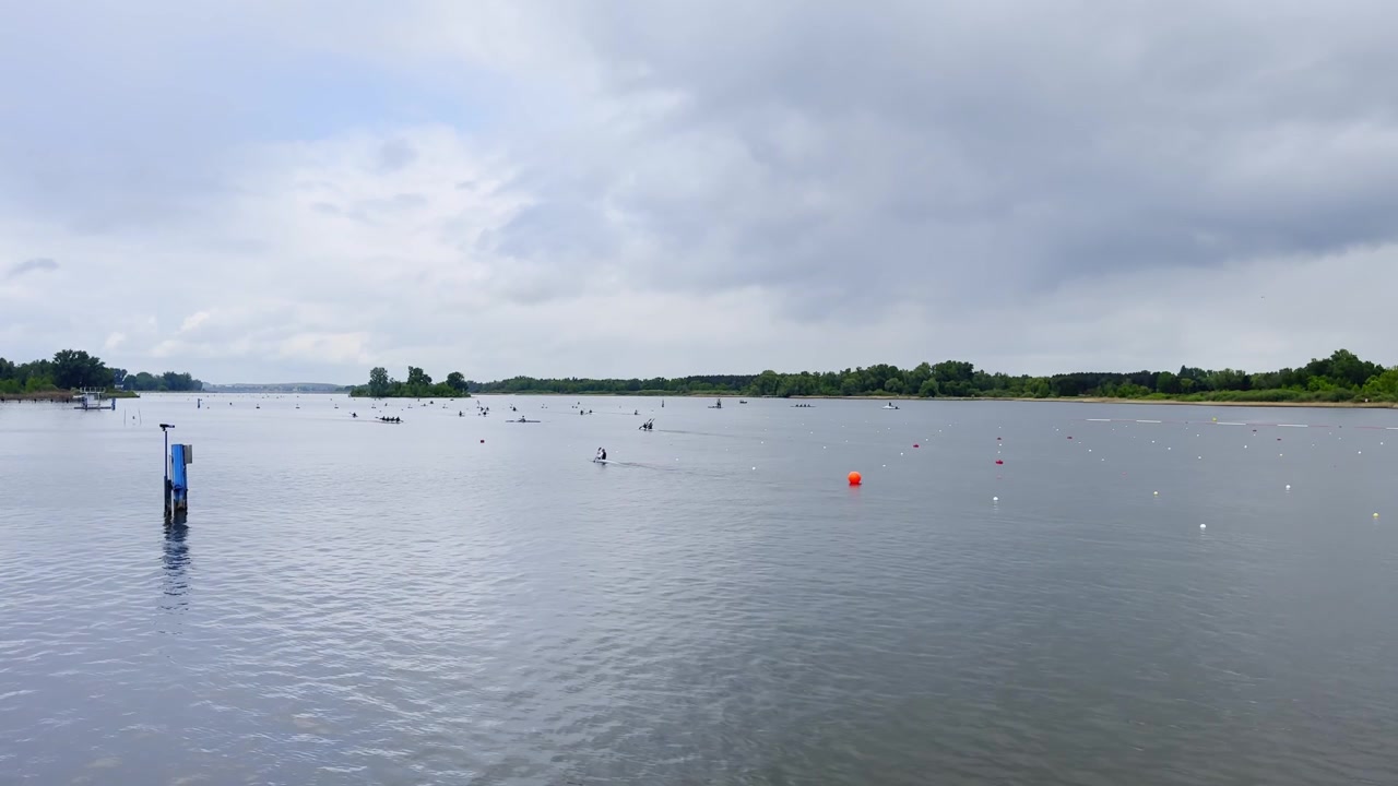 Wide view of a Brandenburg canoe sprint lake venue, with distant paddlers, lane buoys, calm grey water, shoreline trees and overcast race-day sky.