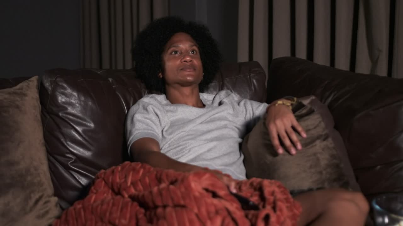 African American man reclining on leather sofa in living room, pressing remote to start show