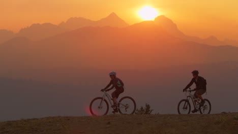 two mountain bikers pull to a stop on a hillside at sunset