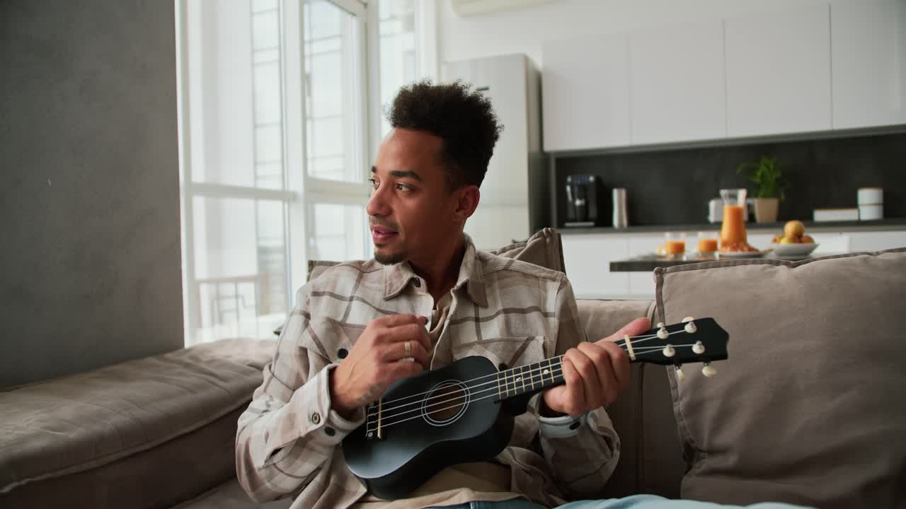 A calm and happy man with Black skin color a young brunette with stubble plays a small musical stringed instrument the ukulele while sitting on a modern sofa in a cream plaid shirt in a modern apartment