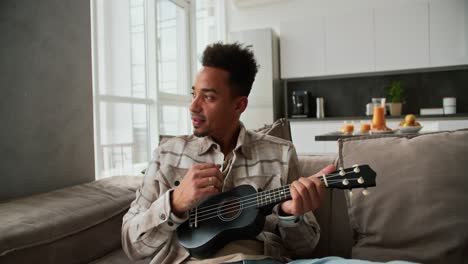 a calm and happy man with black skin color a young brunette with stubble plays a small musical stringed instrument the ukulele while sitting on a modern sofa in a cream plaid shirt in a modern apartment