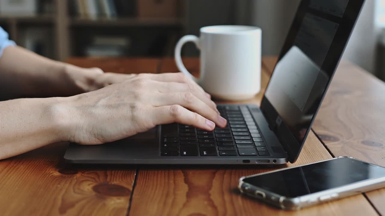 Dita posate che digitano, mani che premono i tasti del laptop a casa, modificando un documento vicino a una tazza e un telefono.