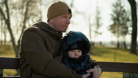 father and son sitting on a bench, with the father resting his head gently on his son's head, with the father holding his son close, with trees in the background