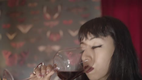 chica atractiva con lápiz labial rojo y piel pálida bebe vino tinto de un vaso grande en una habitación decorada en estilo retro