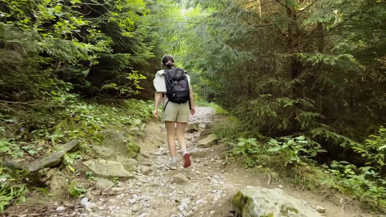 Uma caminhante com uma mochila sobe uma trilha rochosa e íngreme, cercada por uma densa floresta verde, durante uma aventura