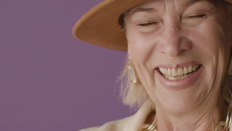 close up view of blonde mature woman with blue eyes dressed in jacket and hat posing wiping tears and smiling at camera on purple background