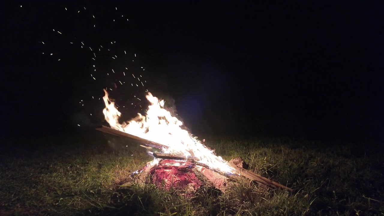 Cozy bonfire blazing on a grassy meadow at night, casting warm firelight and glowing sparks into the darkness. Perfect for camping, hiking, wilderness and outdoor adventure themes.
