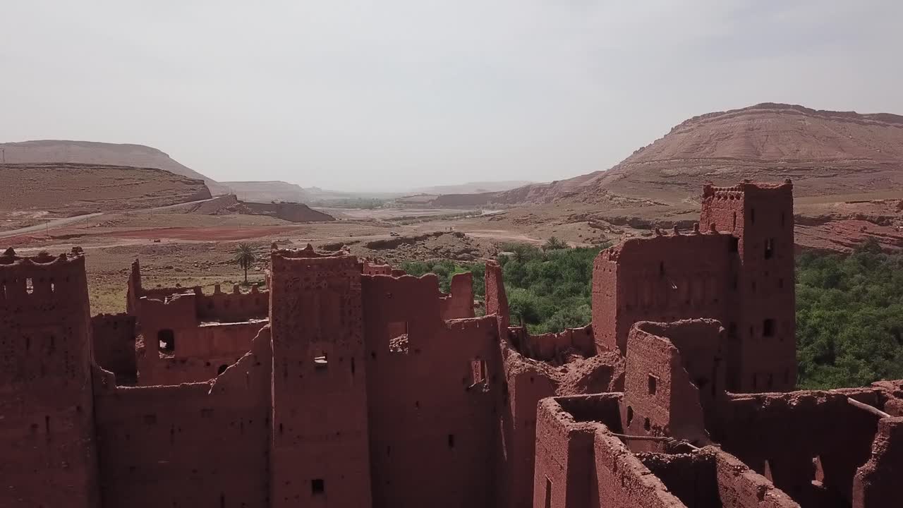 Vue cinématographique d'anciennes ruines de kasbah marocaine entourées de montagnes désertiques et de verdure d'oasis. Architecture traditionnelle en terre dans un cadre historique nord-africain, idéale pour le voyage et le patrimoine.