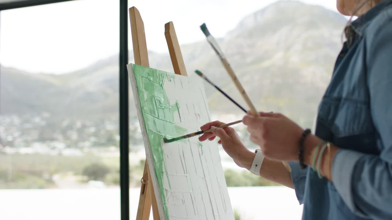 Painter is dipping brush into green paint, applying strokes to underpaint canvas with valley view